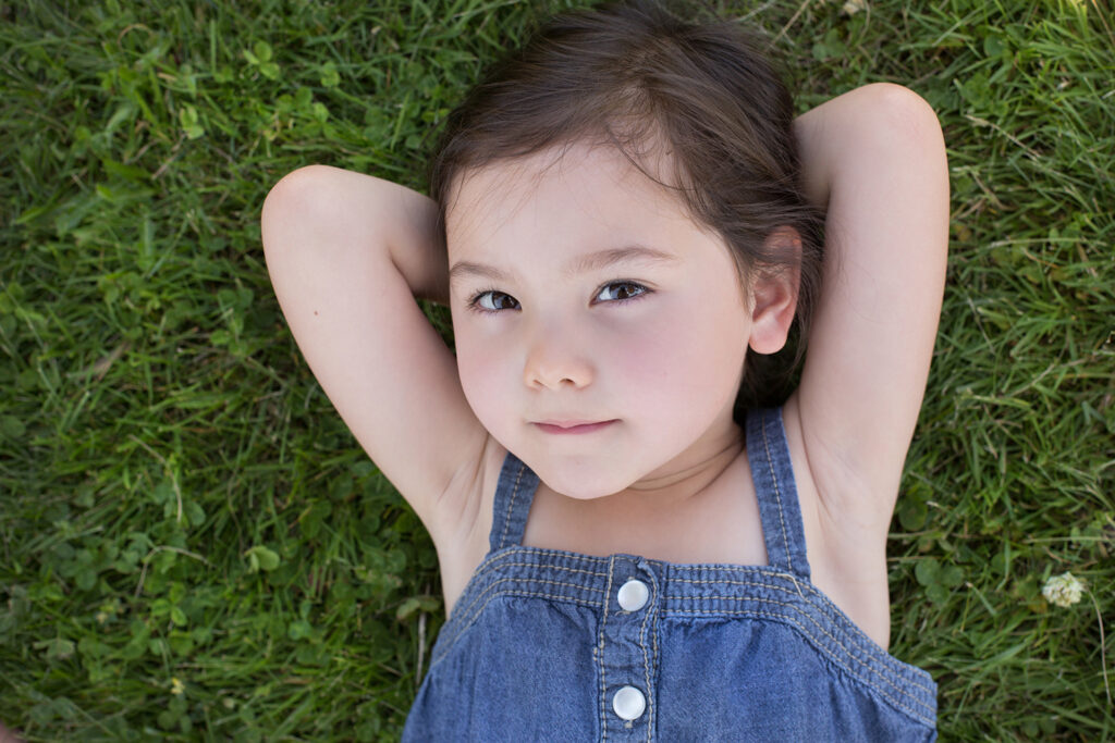 A toddler girl is laying in a grassy field at Heritage Park in Kirkland, Washington. She has her hands crossed behind her head and is looking up at the camera with a serious expression on her face.