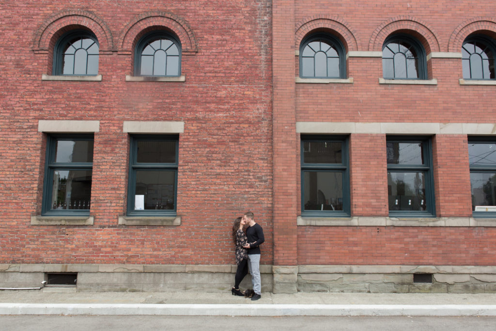 A brunette woman and brunette man kiss in front of a large brick building in an area of Seattle, Washington called Georgetown.