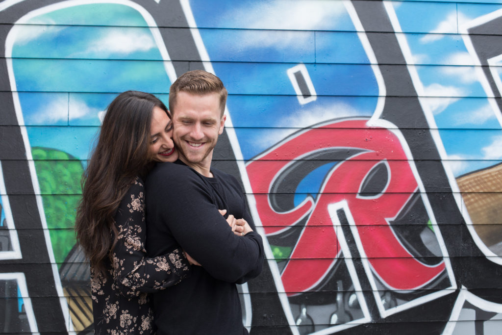 A brunette woman and brunette man snuggle in front of a large wall of graffiti in an area of Seattle, Washington called Georgetown.