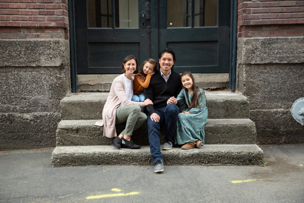 A family of four is cuddled together while sitting on concrete stairs in front of an old brick building in an area of Seattle, Washington called Georgetown. The woman is wearing a long pink cardigan, a white ruffled shirt and green pants, and is snuggling with her younger daughter while holding her husband's hand. The man is wearing a blue button down shirt and jeans. The younger daughter is wearing a yellow long sleeved shirt and blue jean skirt and is resting her head on her dad's shoulder. The older daughter is holding hands with her dad and is wearing a long teal dress with pink flowers.