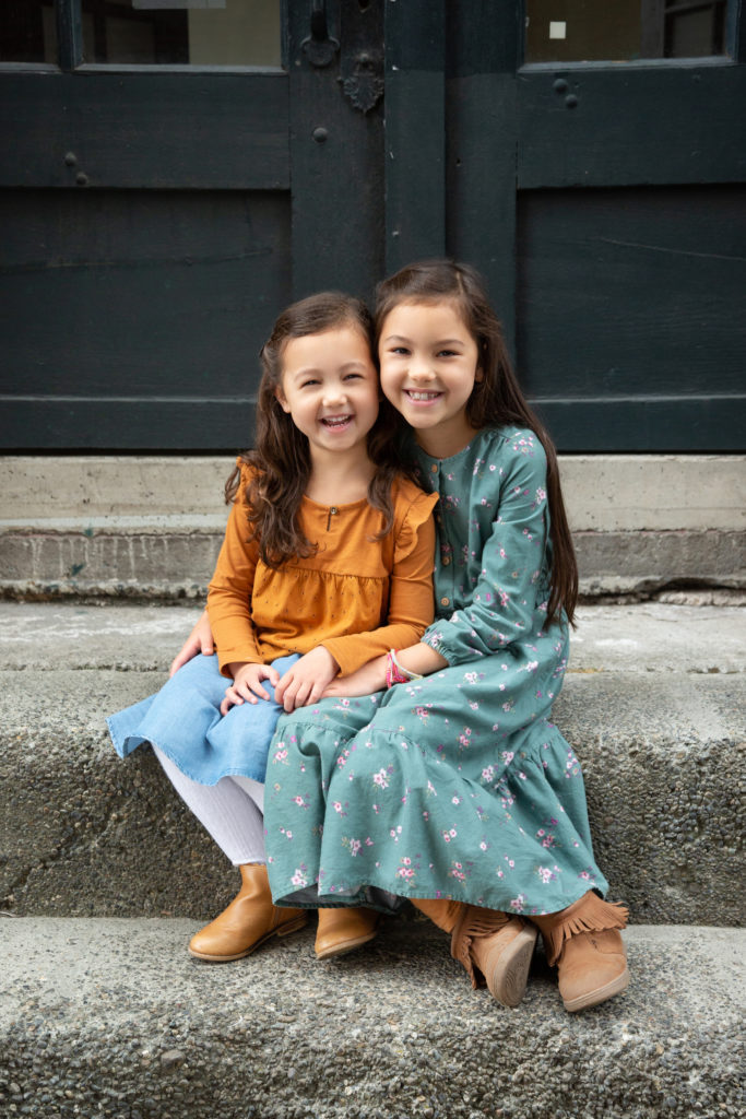 Two sisters cuddle while sitting next to each other on concrete stairs. They're in front of a dark green door of an old building in an area of Seattle, Washington called Georgetown. The younger sister is wearing a yellow long sleeved shirt and blue jean skirt. The older sister is wearing a long teal dress with pink flowers, yellow leggings, and brown boots with fringe.