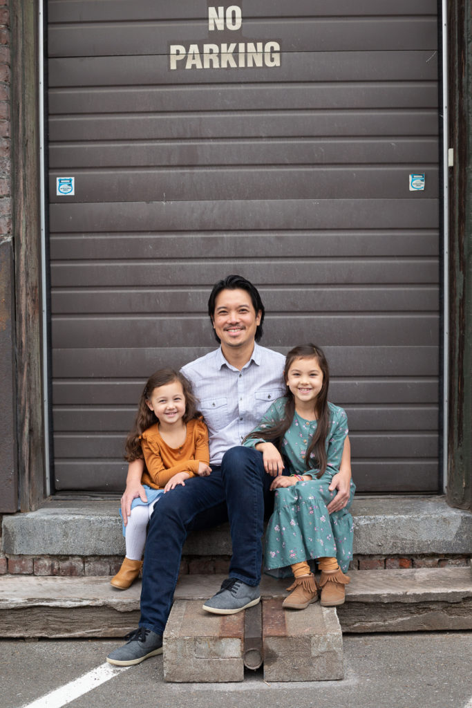 A man wearing a blue button down shirt and jeans cuddles with his two daughters, one is wearing a yellow long sleeved shirt and blue jean skirt and the other is wearing a long teal dress with pink flowers and yellow leggings. They're sitting on a stoop in front of the metal garage door of an old brick building in an area of Seattle, Washington called Georgetown.