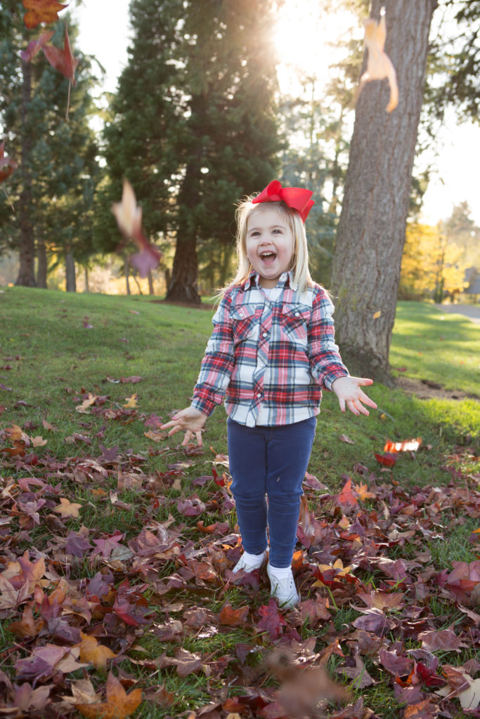 A toddler girl is laughing while throwing red leaves in the air under a tree at Medina Park in Washington state in the fall. Red leaves are scattered all around her.