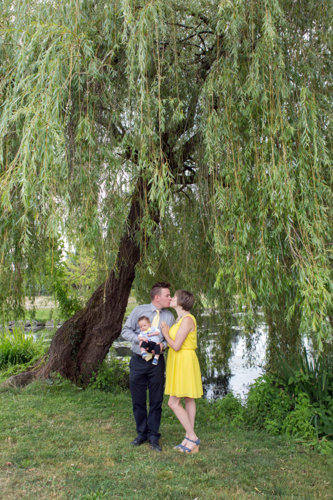 A mom, dad & their newborn baby are cuddled under a willow tree in front of a pond at Medina Park in Washington state in the summer. Mom & dad are kissing while dad holds the baby.