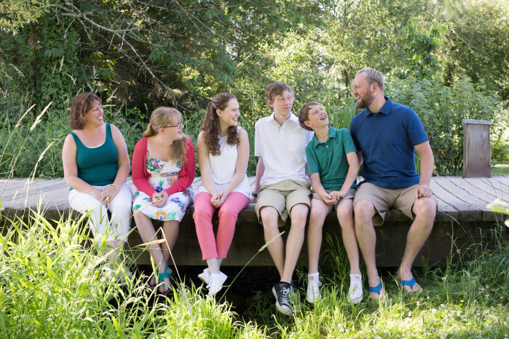 A family of six is sitting together on a wooden bridge at Medina Park in Washington state in the summer. The family is wearing vibrant outfits with blue, teal, pink & white.