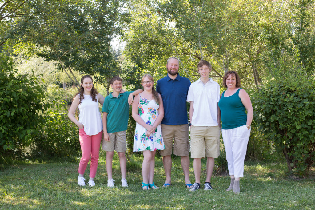 A family of six is standing together in front of green trees and bushes at Medina Park in Washington state in the summer. The family is wearing vibrant outfits with blue, teal, pink & white.