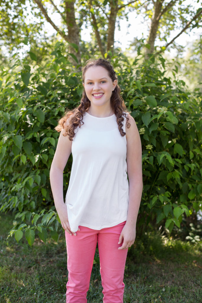 A teenage girl with long, curly red hair wearing a white sleeveless shirt and bubble gum pink pants is standing with her hand in her pocket in front of a green plant at Medina Park in Washington state.