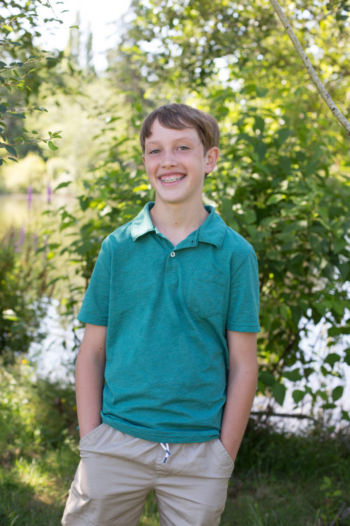 A tween boy with short blonde hair is wearing a teal polo shirt and beige khaki shorts. He is standing with his hands in his pockets in front of a green plant at Medina Park in Washington state.