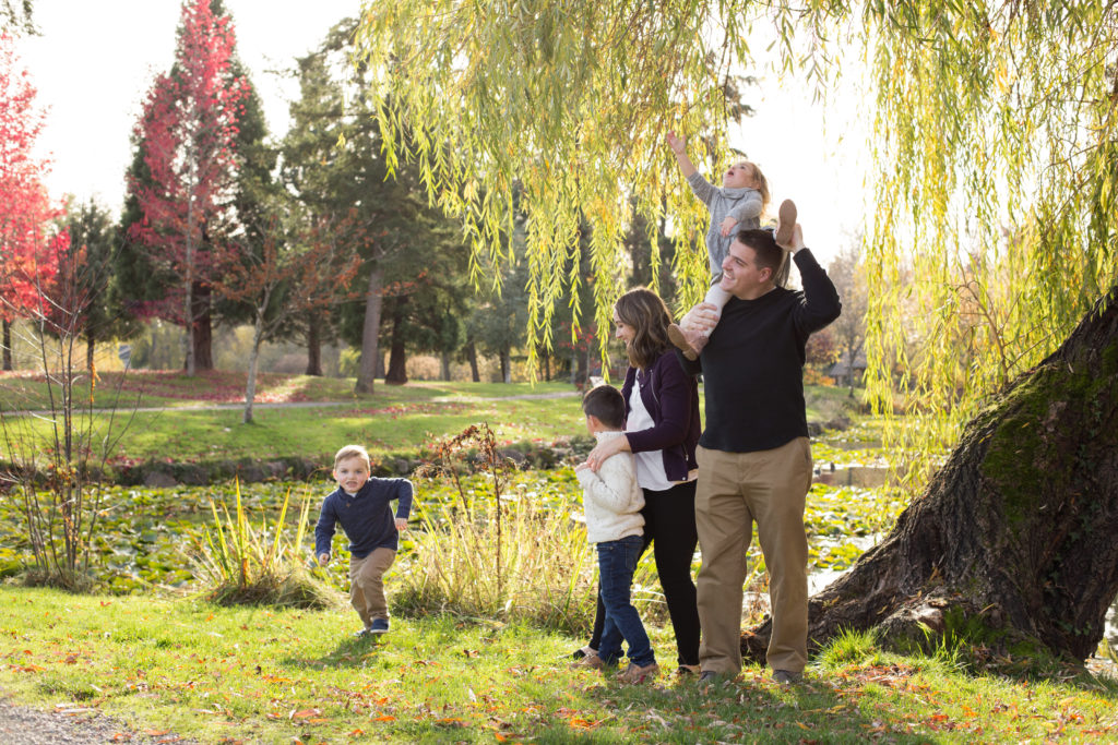 A family of five is playing together while standing under a willow tree in front of a pond at Medina Park in Washington state during fall. The youngest daughter is on her dad's shoulders, reaching up to the leaves on the willow tree. The oldest son is cuddling with mom. The middle son is running towards the camera.