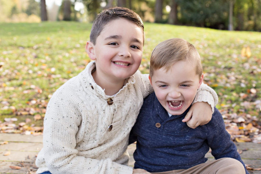 Two brothers laugh and cuddle while sitting next to each other on a wooden dock. They're in front of a grassy hill at Medina Park in Washington state. The younger brother is wearing a blue long sleeved sweater and brown khakis. The older brother is wearing a cream colored sweater.