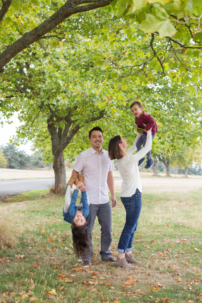 A family of four is standing together under a green tree at Discovery Park in Seattle, Washington. The woman is holding her toddler daughter in the air while looking at her. The dad is holding his daughter's hand while she swings upside down from his arm. 