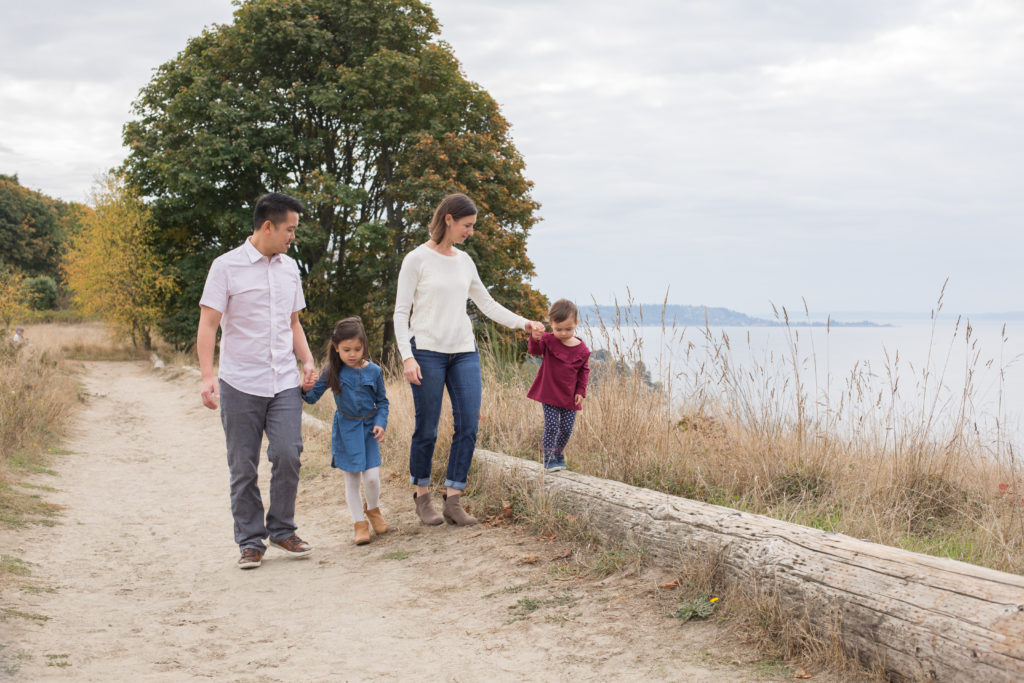 A family of four is walking together at Discovery Park in Seattle, Washington. The mom is holding her toddler's hand while she walks balancing on a log. The dad is holding the other daughter's hand while looking at her. You can see trees with green & yellow leaves in the background, as well as the water of the Puget Sound.
