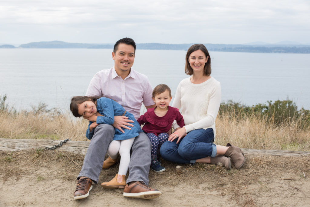 A dad, mom and two daughters are sitting together on a log at Discovery Park in Seattle, Washington. The Puget Sound is visible behind them.