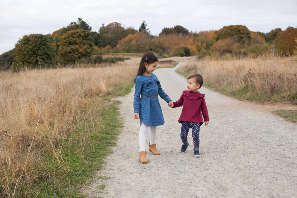 Two young sisters are walking together on a gravel path at Discovery Park in Seattle, Washington. They are holding hands and looking at each other. Trees with green, yellow and orange leaves are in the background. Tall beige & green grass are on either side of the gravel pathway.