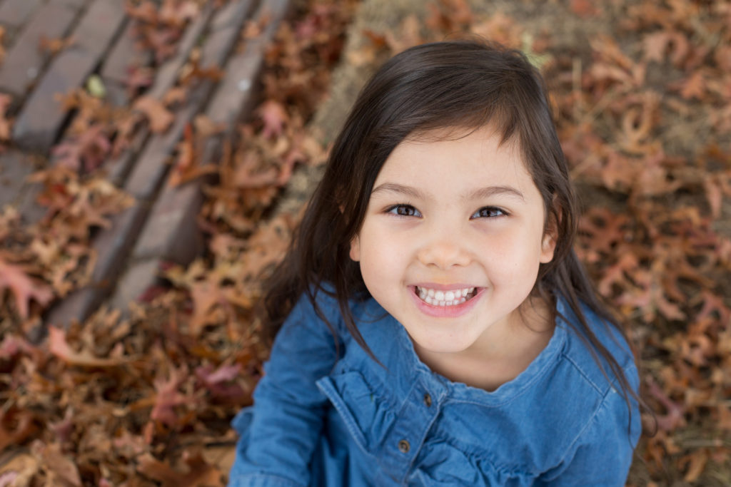 A little girl wearing a blue dress is looking up at the camera and smiling. She's sitting in a pile of orange leaves at Discovery Park in Seattle, Washington.