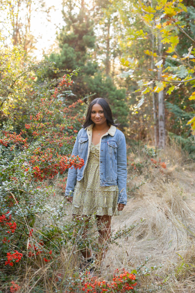 A high school senior girl is standing in a field of tall, cream colored grass in the fall at Discovery Park in Seattle, Washington. Her hands are by her side and she is smiling while looking at the camera. Vibrant red berries and green trees are all around her.