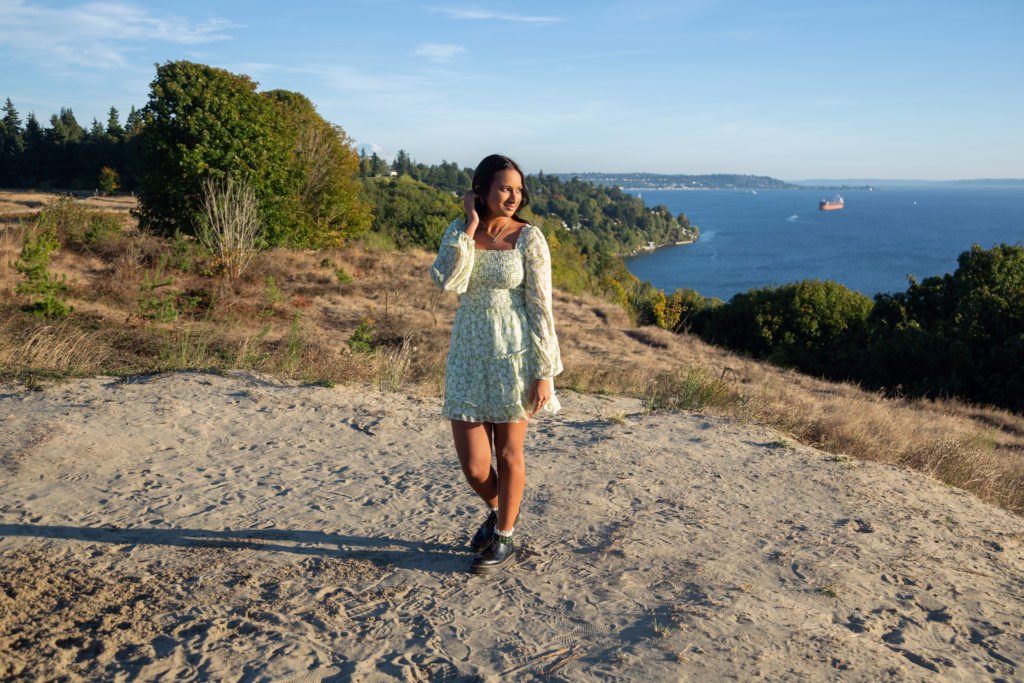 A high school senior girl is standing on the sandy bluff at Discovery Park in Seattle, Washington. Her hand is in her hair and she is smiling slightly while looking away from the camera. You can see the Puget Sound and Mt Rainier in the background. Green trees and hillsides are also visible.