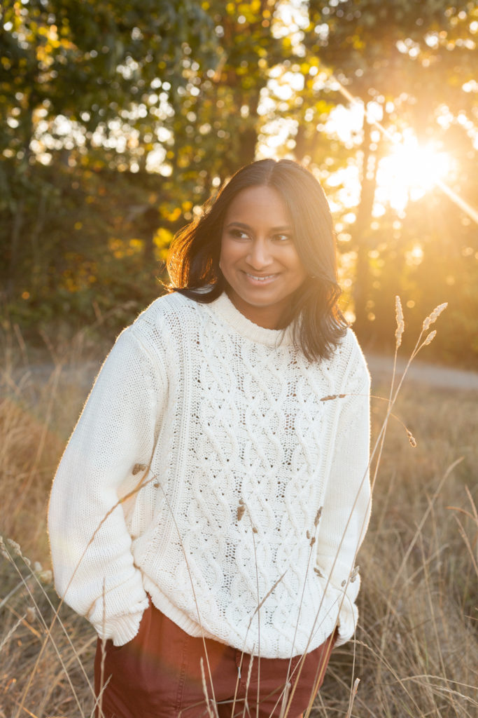 A high school senior girl is standing in a field at sunset during golden hour at Discovery Park in Seattle, Washington. Her hands are in her pockets and she is smiling while looking away from the camera.