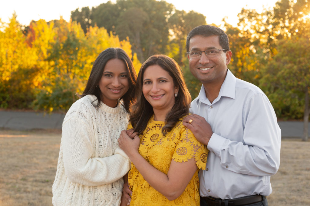 A high school senior girl is standing with her mom and dad in front of vibrant yellow leaves at sunset during golden hour at Discovery Park in Seattle, Washington. They are cuddled together and smiling at the camera.