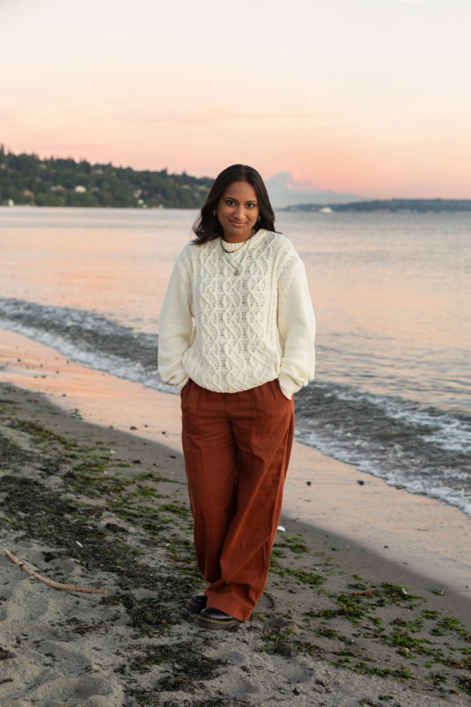 A high school senior girl is standing on the beach at sunset during golden hour at Discovery Park in Seattle, Washington. Her hands are in her pockets and she looking at the camera. The Puget Sound and Mt Rainier are in the background.