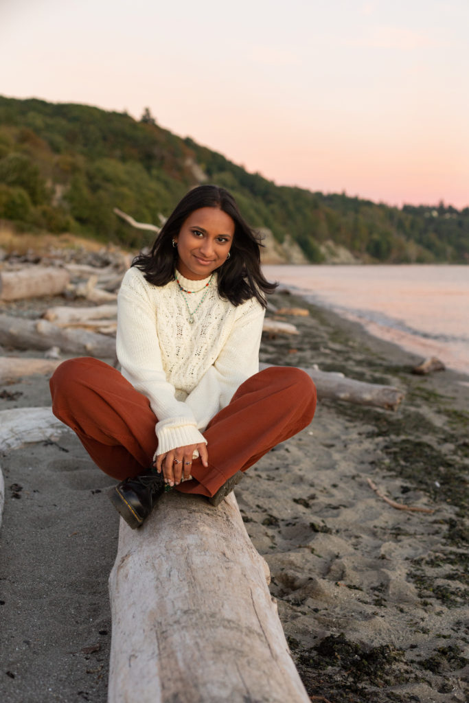 A high school senior girl is sitting on a piece of driftwood at the beach at sunset during golden hour at Discovery Park in Seattle, Washington. Her legs are crossed and her hands are by her feet. The Puget Sound is in the background along with a hillside of trees with green and orange leaves.