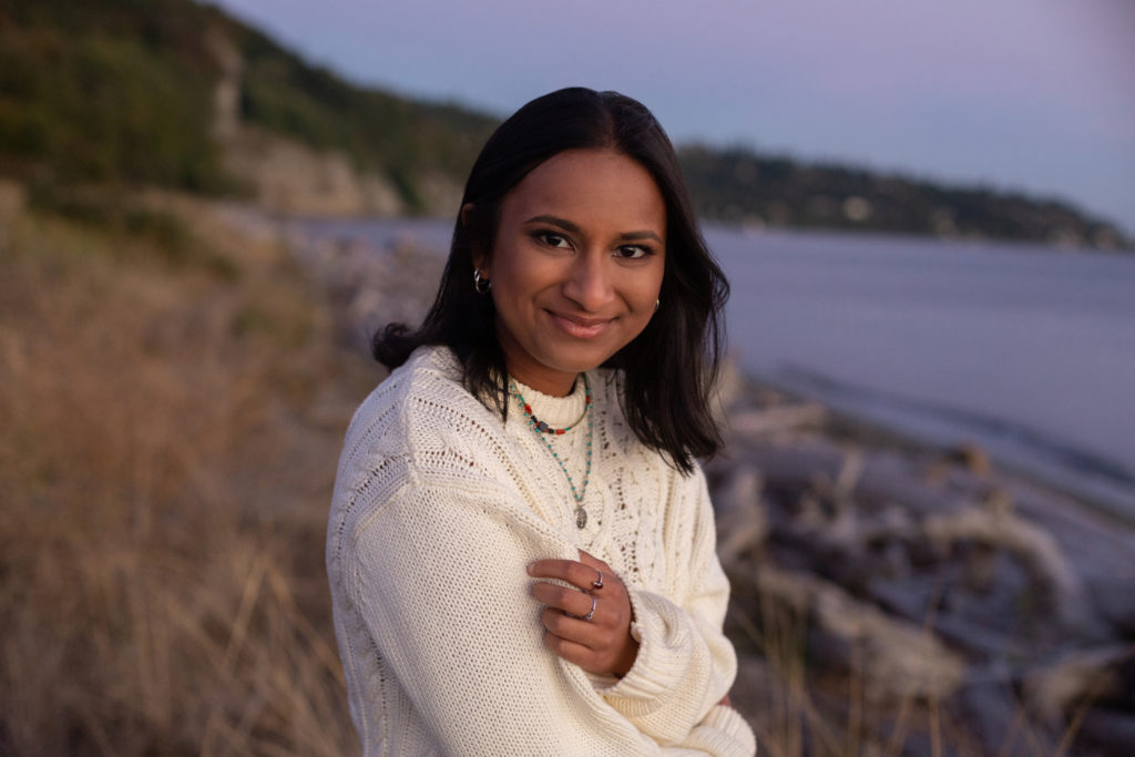 A high school senior girl is standing on a beach at sunset during golden hour at Discovery Park in Seattle, Washington. She is wearing a cream colored sweater along with necklaces and rings. Her arms are crossed and her hand is resting on her opposite arm. She is smiling while looking the camera.