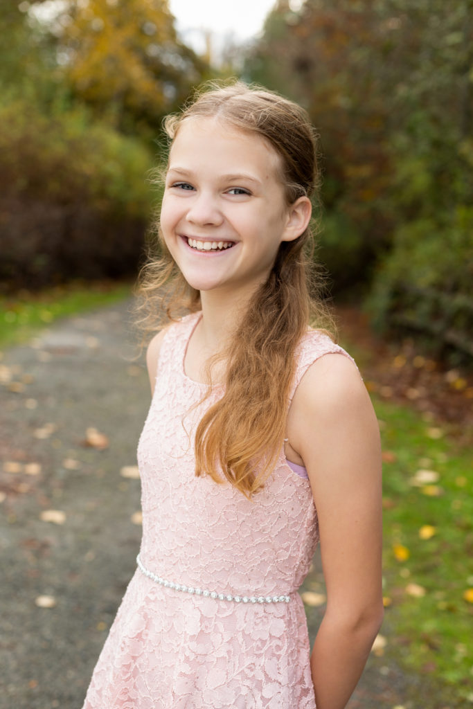 A middle school aged girl is standing in front of a tree lined pathway with yellow leaves on the ground at Luther Burbank Park on Mercer Island in Washington state. She is wearing a pink sleeveless dress. She is smiling while looking at the camera.