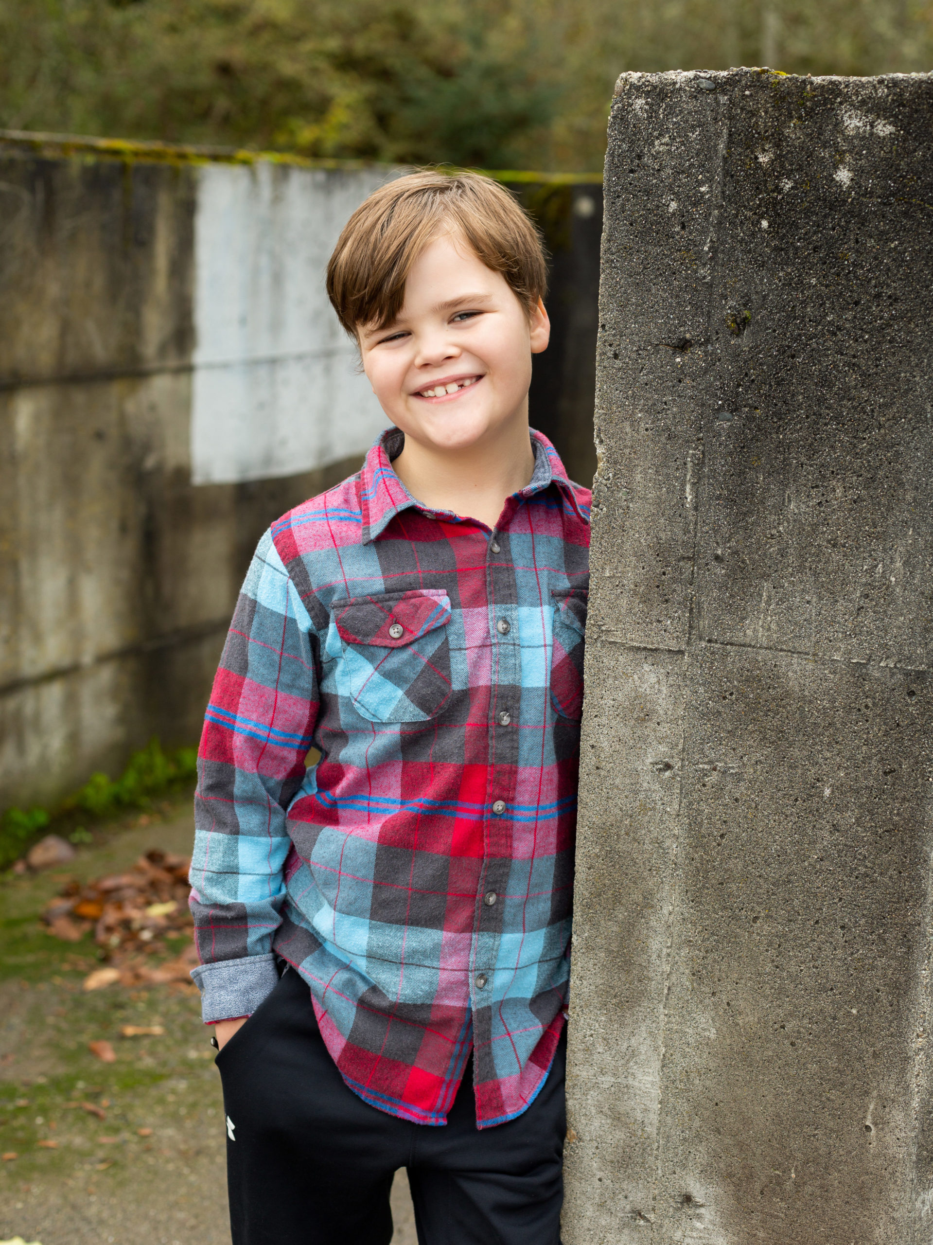 An elementary school aged boy is leaning against a concrete wall of an old building at Luther Burbank Park on Mercer Island in Washington state. The boy is wearing a blue and red flannel shirt. He's got his hand in his pocket and he is smiling at the camera.