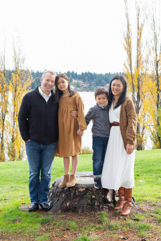 A mom, dad, 5 year old son and 8 year old daughter are standing together holding hands at Luther Burbank Park on Mercer Island near Seattle, WA. They're on a grassy hill lined with green trees and Lake Washington is behind them. The family is smiling at the camera.