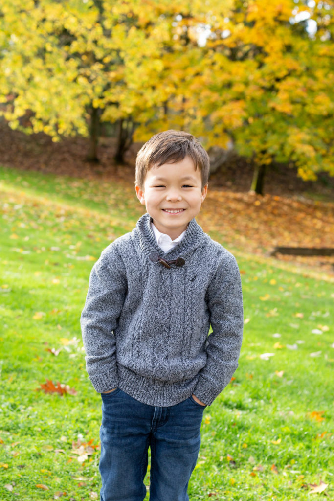 A small boy is standing in front of a tree lined hill on Mercer Island in Washington state. It's fall and the leaves are vibrant shades of yellow, orange and green. He is wearing a gray sweater with blue jeans. He is smiling & looking at the camera.