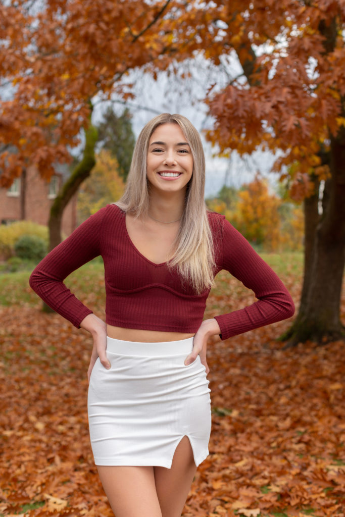A high school senior girl is standing with her hands on her hips in front of a tree with red leaves at Luther Burbank Park on Mercer Island in Washington state. She is wearing a maroon long sleeve shirt & white skirt. She is smiling & looking at the camera.