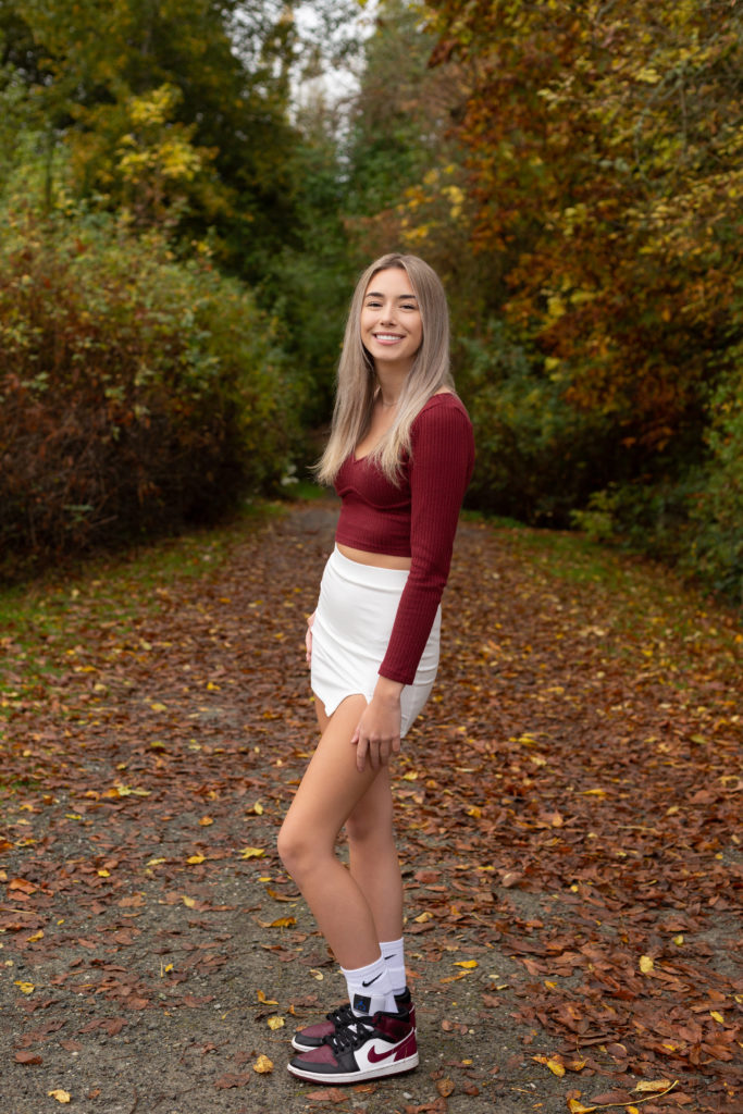 A high school senior girl is standing in front of a tree lined pathway with red leaves on the ground at Luther Burbank Park on Mercer Island in Washington state. She is wearing a maroon long sleeve shirt & white skirt. She is smiling & looking at the camera.