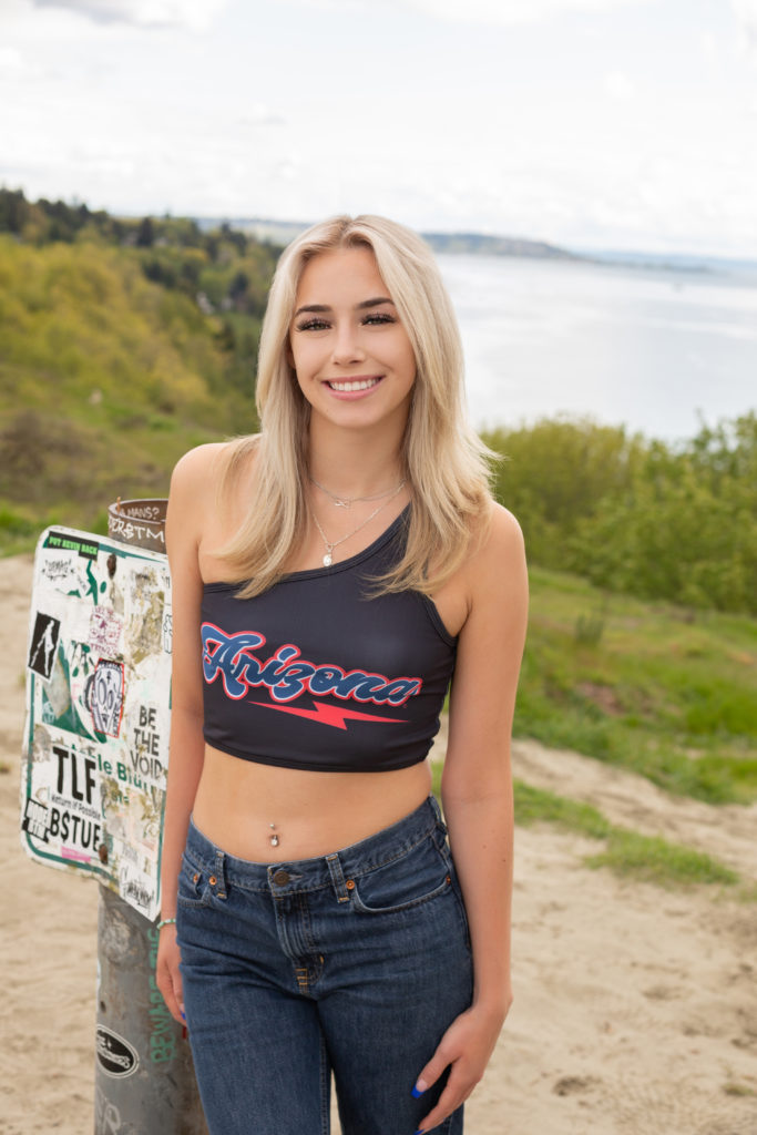 A high school senior girl is standing on the sandy bluff at Discovery Park in Seattle, Washington. She is leaning against a sign covered by stickers while smiling at the camera. You can see the Puget Sound 
 in the background. Green trees and hillsides are also visible.