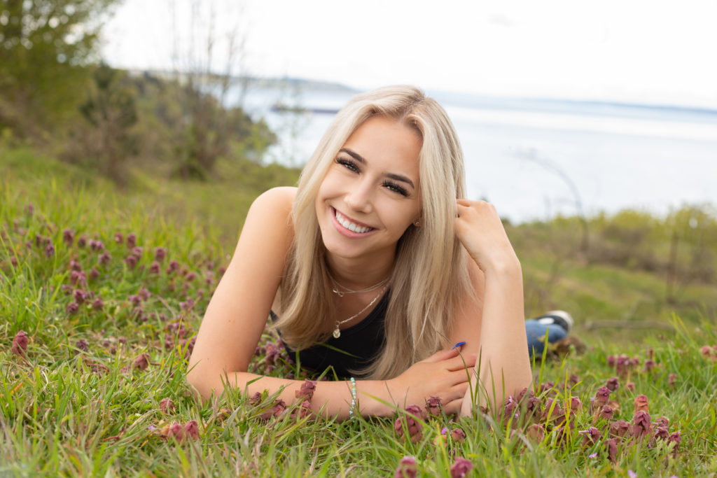 A high school senior girl is laying on her stomach with her hand in her hair on a grassy and purple flower-filled hillside at Discovery Park in Seattle, Washington. She is smiling at the camera and the waters of Puget Sound are behind her.