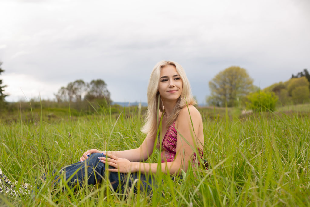 A high school senior girl is sitting in a grassy field at Discovery Park in Seattle, Washington. She is smiling while looking away from the camera. Green trees and grass are behind her.