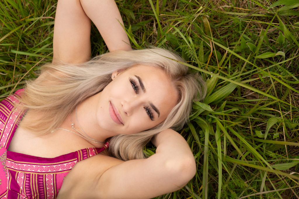 A high school senior girl is laying down in a grassy field, her head resting on her arms crossed behind them at Discovery Park in Seattle, Washington. The photographer is standing above her, so she is smiling while looking up at the camera.