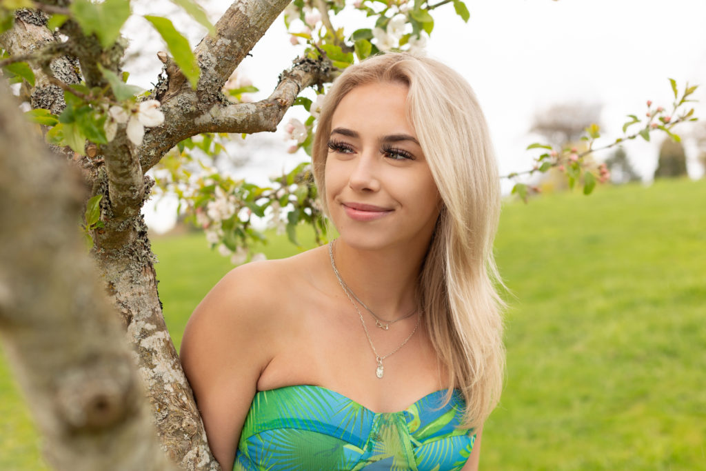 A high school senior girl is standing next to a tree with small pink blossoms at Discovery Park in Seattle, Washington. She is smiling while looking away from the camera. Green trees and grass are behind her.