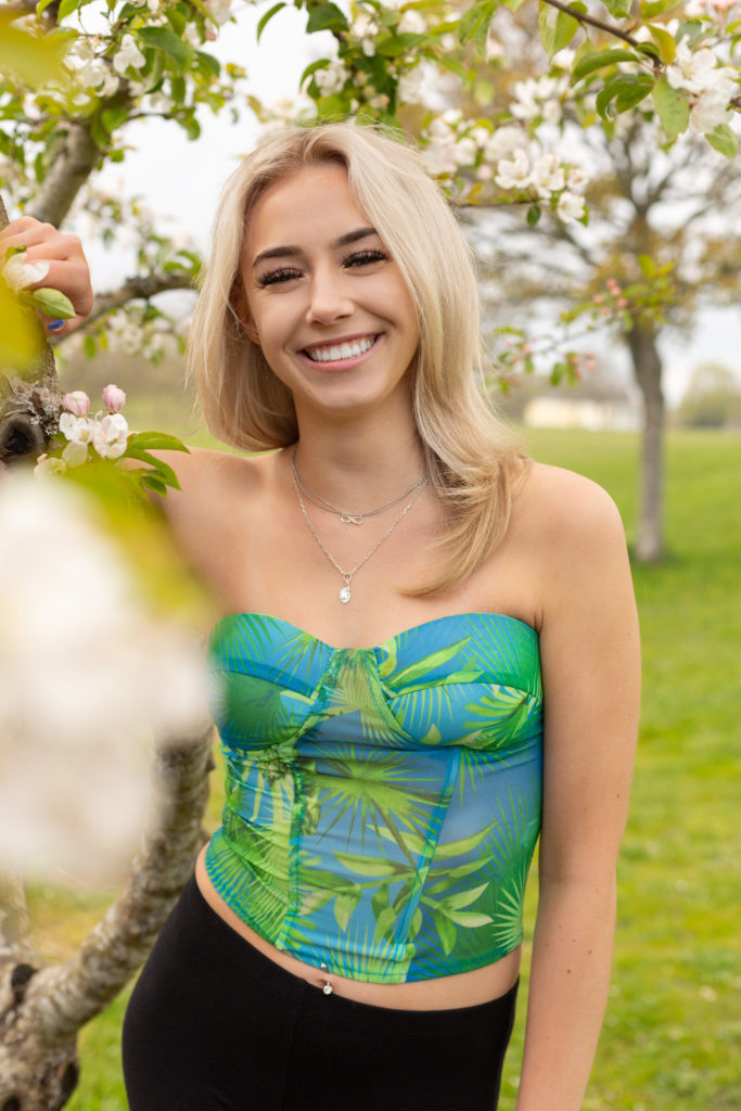 A high school senior girl is standing next to a tree with small pink blossoms at Discovery Park in Seattle, Washington. She is smiling at the camera. Green trees and grass are behind her.