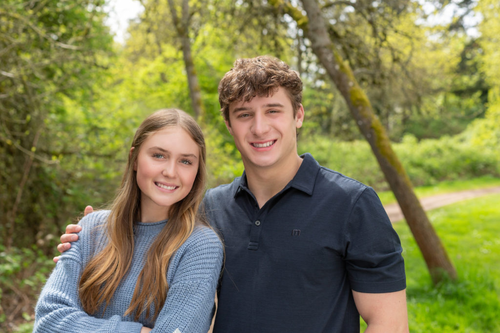 A teenage brother & sister are standing together at Luther Burbank Park on Mercer Island near Seattle, WA. They're on a gravel path lined with green trees. The brother has his arm around his sister & they're smiling at the camera.