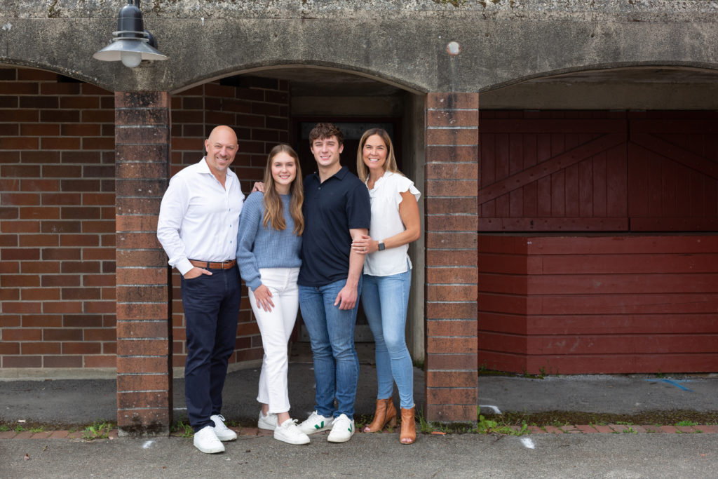 A mom, dad, teenage son & teenage daughter are standing together at Luther Burbank Park on Mercer Island near Seattle, WA. They're under a brick & concrete archway with a brick building behind them. The family is smiling and looking at the camera.