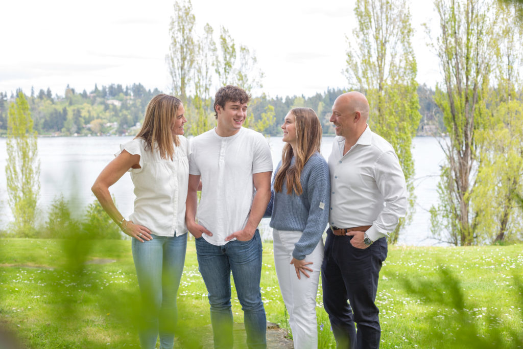 A mom, dad, teenage son & teenage daughter are standing together at Luther Burbank Park on Mercer Island near Seattle, WA. They're on a gravel path lined with green trees and Lake Washington is behind them. The family is smiling and laughing while looking at each other.