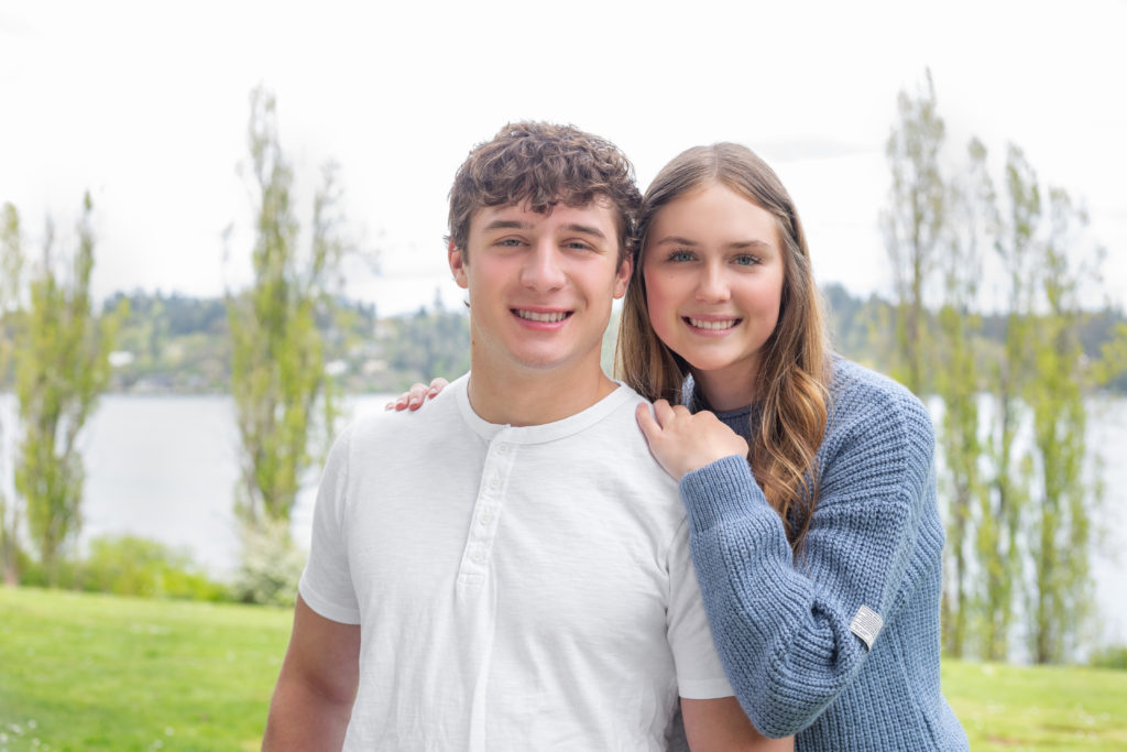 A teenage brother & sister are standing together at Luther Burbank Park on Mercer Island near Seattle, WA. They're on a gravel path lined with green trees and Lake Washington is behind them. The sister has her arms on her brother's shoulders & they're smiling at the camera.