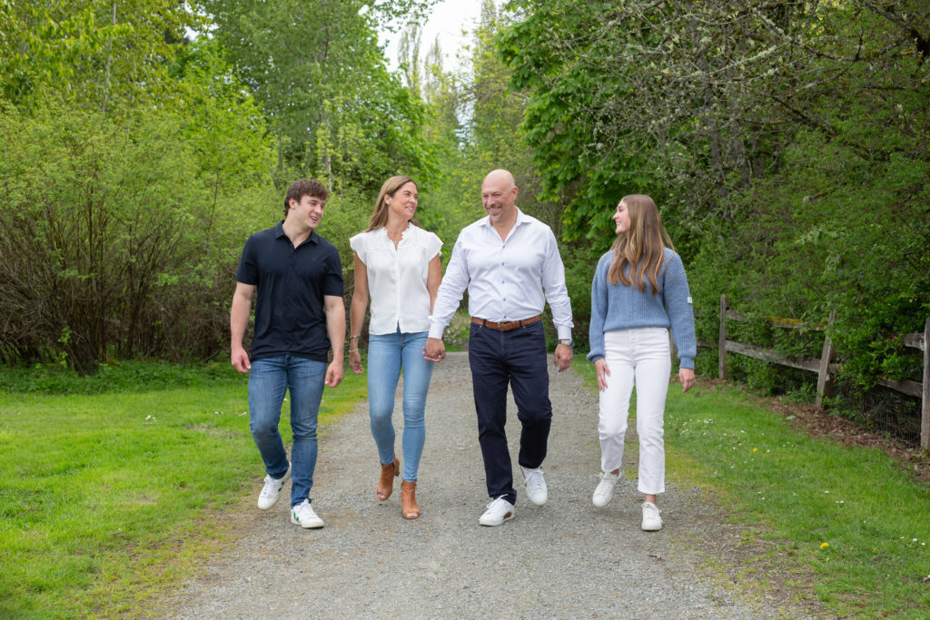 A mom, dad, teenage son & teenage daughter are walking together at Luther Burbank Park on Mercer Island near Seattle, WA. They're on a gravel path lined with green trees and an old brown fence. The family is smiling and laughing while looking at each other.