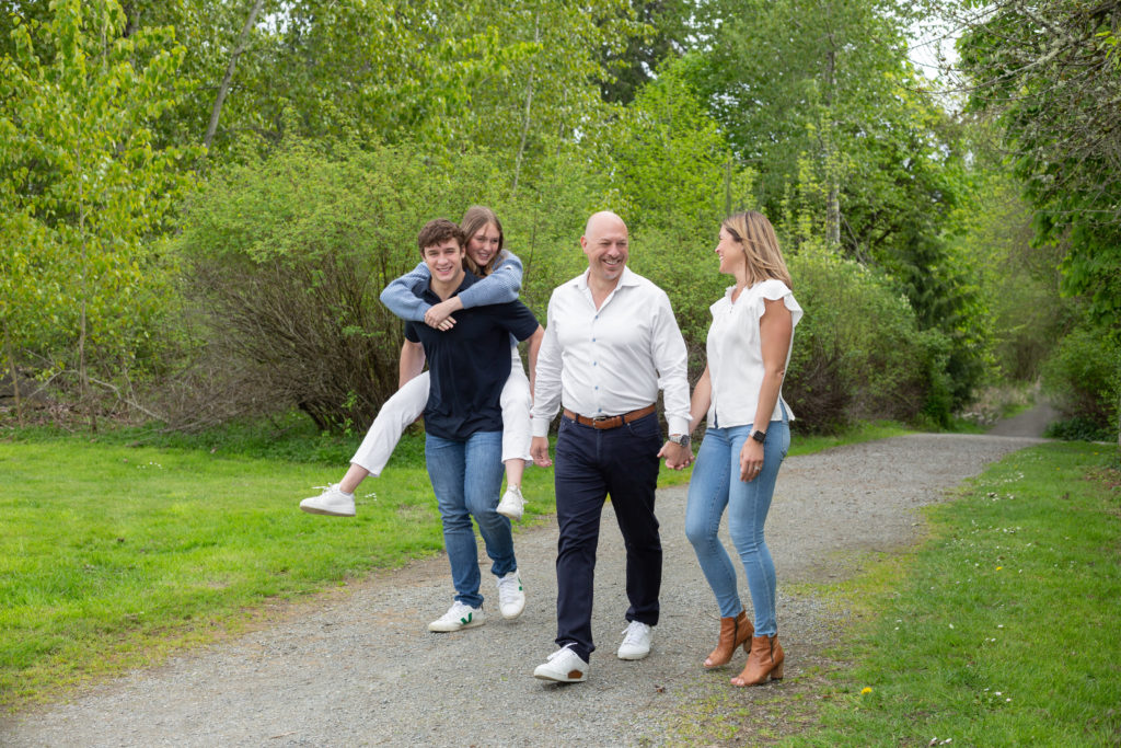 A mom, dad, teenage son & teenage daughter are walking together at Luther Burbank Park on Mercer Island near Seattle, WA. They're on a gravel path lined with green trees. The brother is giving his sister a piggy back ride while their parents hold each other's hands. The family is smiling and laughing while looking at each other.
