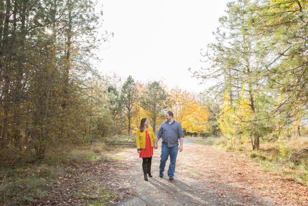 A woman and man are holding hands while walking together down a gravel path at Discovery Park in Seattle, Washington. It's fall and there are yellow, orange and green leaves on the trees surrounding them.