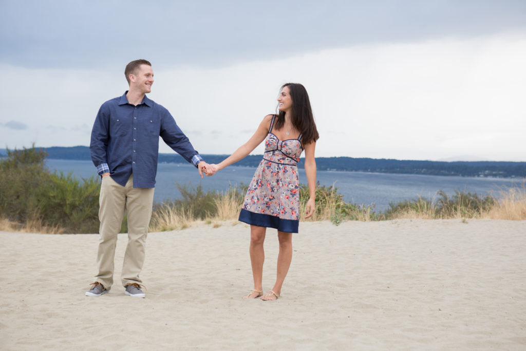 A man and woman are standing and holding hands on the sandy bluff at Discovery Park in Seattle, Washington. They are looking at each other smiling and laughing. The Puget Sound is in the background.