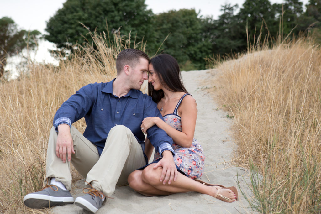 A man and woman are sitting on a sandy path with tall grass on either side of them at Discovery Park in Seattle, Washington. The man is sitting with his legs bent and the woman is cuddled up next to him holding onto his arm. They are looking at each other with their foreheads touching.