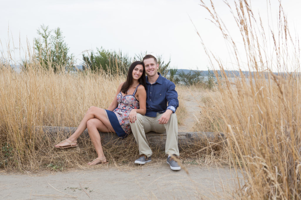 A woman and man are sitting together on a log at Discovery Park in Seattle, Washington. Tall dried up grass is surrounding them and the Puget Sound is visible behind them.