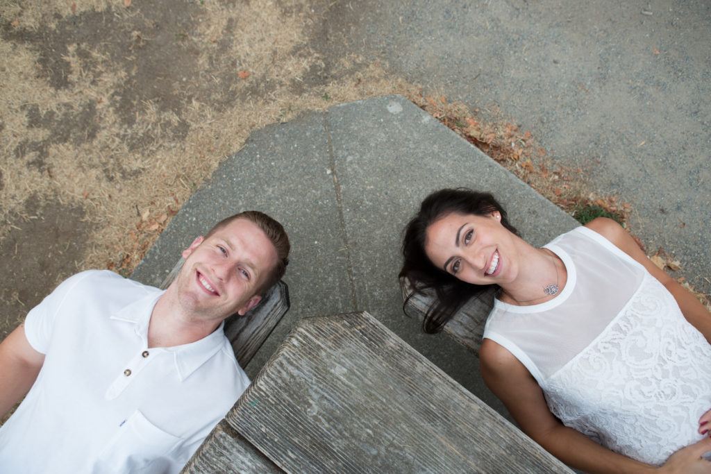 A woman and man are laying down next to each other on separate benches at a picnic table at Discovery Park in Seattle, Washington. The photographer is standing on the table photographing downward. The couple is smiling while looking up at the camera.