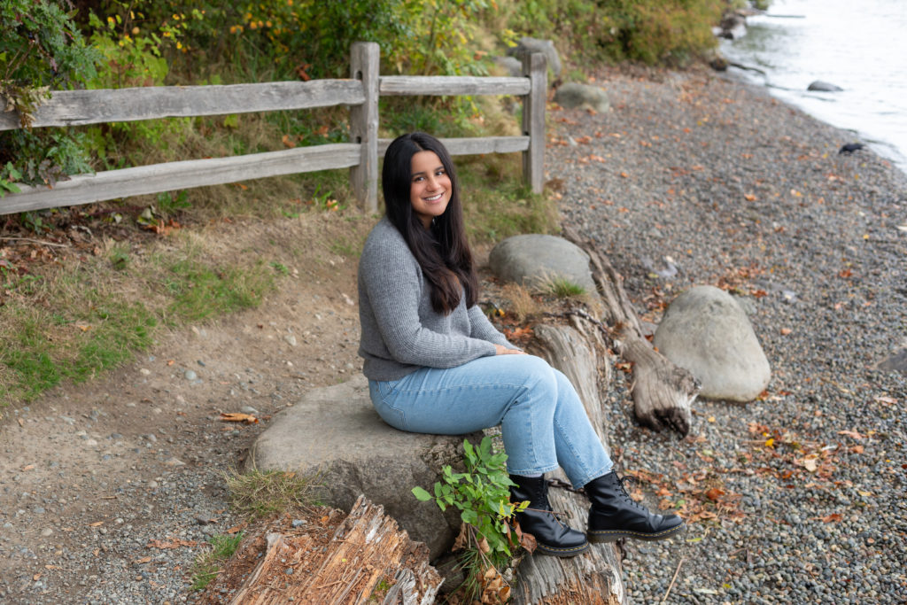 A high school senior girl is sitting on a rock by the shoreline at Luther Burbank Park on Mercer Island in Washington state. There's an old fence and trees behind her and you can see some of Lake Washington next to her. She is wearing a gray sweater, blue jeans and black boots. She is smiling & looking at the camera.